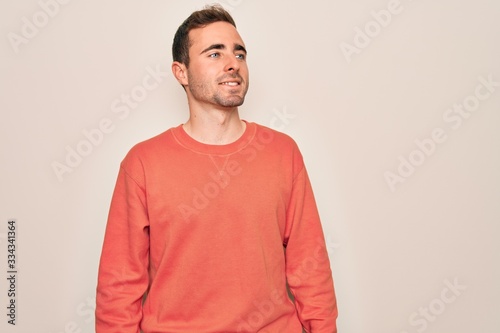Young handsome man with blue eyes wearing casual sweater standing over white background looking away to side with smile on face, natural expression. Laughing confident.