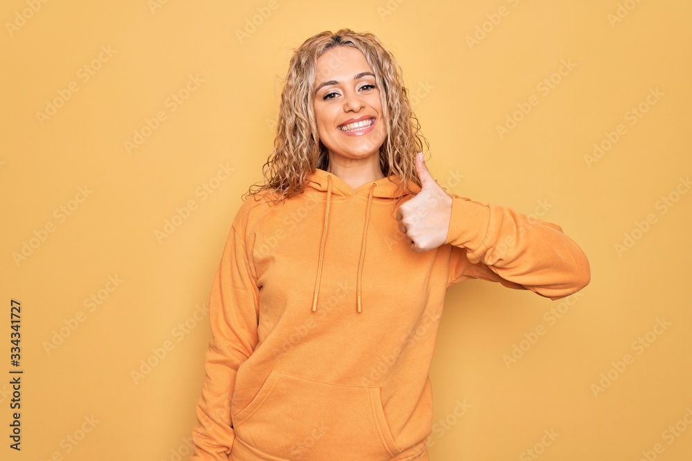 Young beautiful blonde sporty woman wearing casual sweatshirt over yellow background doing happy thumbs up gesture with hand. Approving expression looking at the camera showing success. Young beautiful blonde sporty woman wearing casual sweatshirt over yellow background doing happy thumbs up gesture with hand. Approving expression looking at the camera showing success.