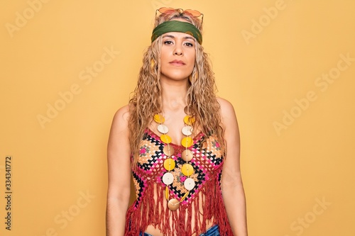 Beautiful blonde hippie woman wearing sunglasses and accessories over yellow background Relaxed with serious expression on face. Simple and natural looking at the camera.