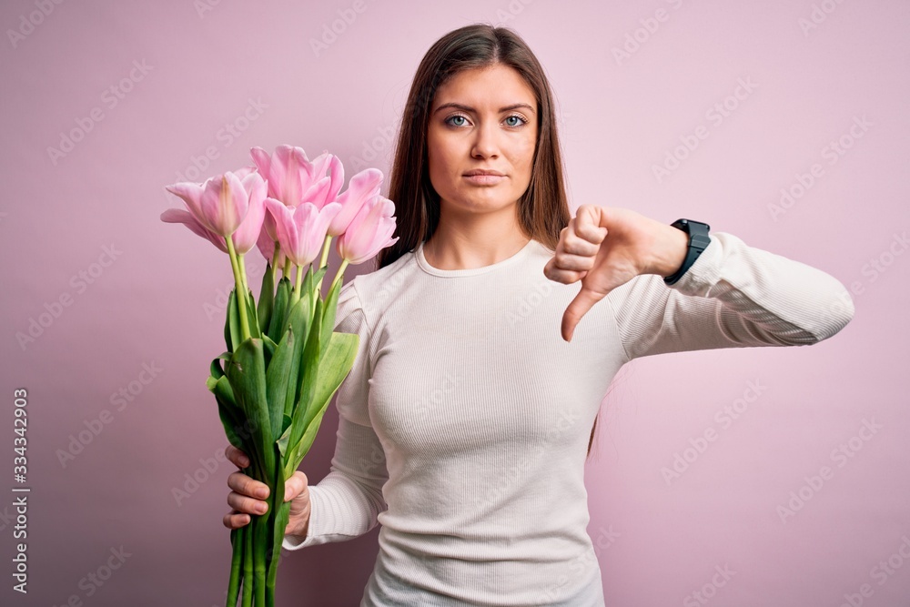 Young beautiful romantic woman with blue eyes holding beauty bouquet of pink tulips with angry face, negative sign showing dislike with thumbs down, rejection concept Young beautiful romantic woman with blue eyes holding beauty bouquet of pink tulips with angry face, negative sign showing dislike with thumbs down, rejection concept