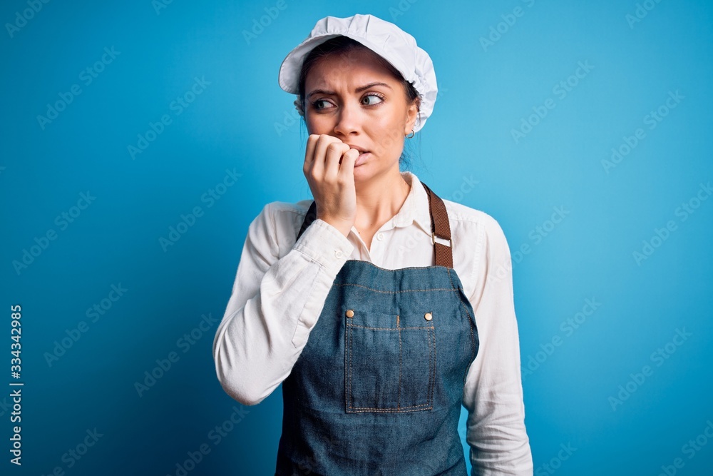 Young beautiful baker woman with blue eyes wearing apron and cap over blue background looking stressed and nervous with hands on mouth biting nails. Anxiety problem. Young beautiful baker woman with blue eyes wearing apron and cap over blue background looking stressed and nervous with hands on mouth biting nails. Anxiety problem.