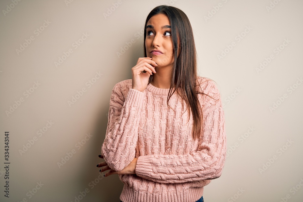 Young beautiful brunette woman wearing casual sweater over isolated white background with hand on chin thinking about question, pensive expression. Smiling with thoughtful face. Doubt concept.