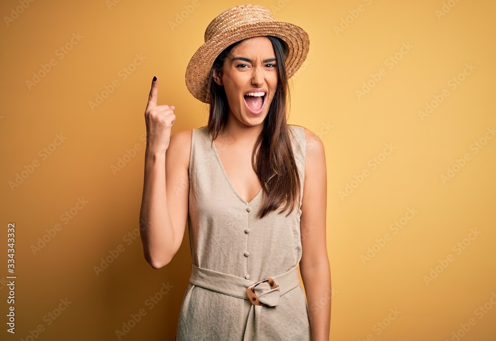 Young beautiful brunette woman on vacation wearing casual dress and hat pointing finger up with successful idea. Exited and happy. Number one. Young beautiful brunette woman on vacation wearing casual dress and hat pointing finger up with successful idea. Exited and happy. Number one.