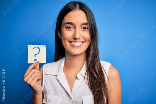 Young beautiful brunette woman holding paper with question mark symbol message with a happy face standing and smiling with a confident smile showing teeth