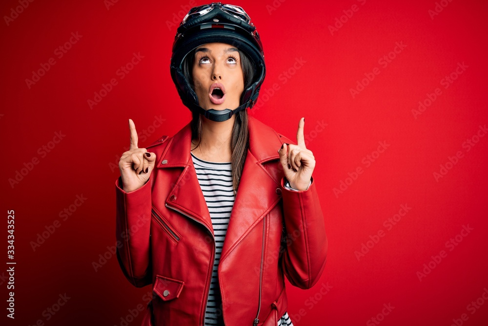 Young beautiful brunette motorcyclist woman wearing motorcycle helmet and red jacket amazed and surprised looking up and pointing with fingers and raised arms. Young beautiful brunette motorcyclist woman wearing motorcycle helmet and red jacket amazed and surprised looking up and pointing with fingers and raised arms.