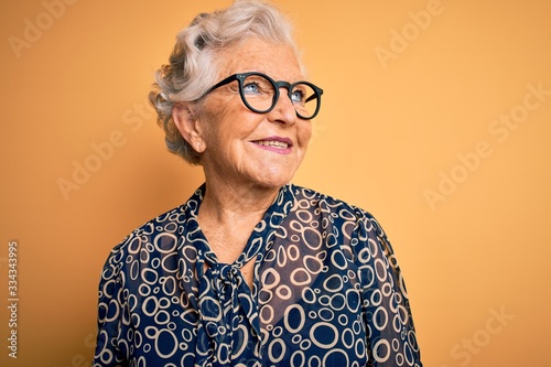Senior beautiful grey-haired woman wearing casual shirt and glasses over yellow background looking away to side with smile on face, natural expression. Laughing confident.