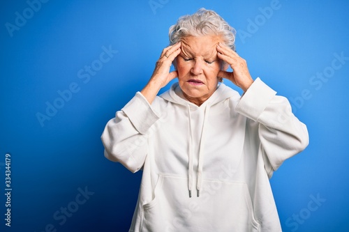 Senior beautiful sporty woman wearing white sweatshirt over isolated blue background suffering from headache desperate and stressed because pain and migraine. Hands on head.