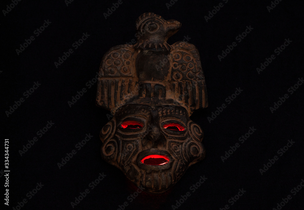 An ancient ceramic pre columbus mask based in American indigenous tribes art iluminated by red light inside over black background 