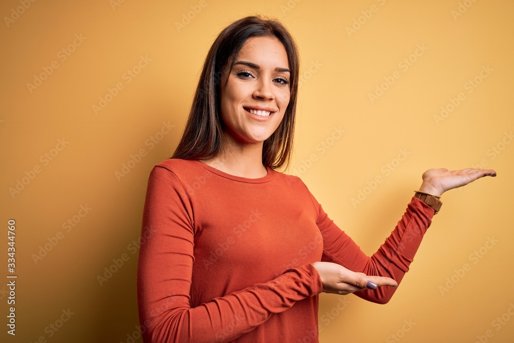 Young beautiful brunette woman wearing casual t-shirt standing over yellow background Inviting to enter smiling natural with open hand Young beautiful brunette woman wearing casual t-shirt standing over yellow background Inviting to enter smiling natural with open hand