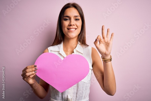 Young beautiful brunette woman holding big pink heart paper celebrating valentine day doing ok sign with fingers, excellent symbol