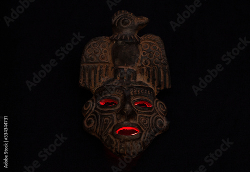 An ancient ceramic pre columbus mask based in American indigenous tribes art iluminated by red light inside over black background 