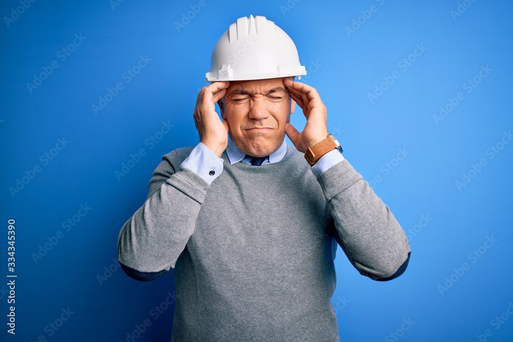 Middle age handsome grey-haired engineer man wearing safety helmet over blue background with hand on head for pain in head because stress. Suffering migraine. Middle age handsome grey-haired engineer man wearing safety helmet over blue background with hand on head for pain in head because stress. Suffering migraine.
