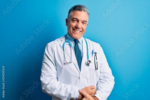 Middle age handsome grey-haired doctor man wearing coat and blue stethoscope with hands together and crossed fingers smiling relaxed and cheerful. Success and optimistic