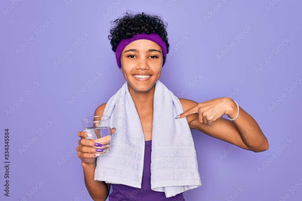 Beautiful african american sporty woman doing exercise wearing towel drinking glass of water with surprise face pointing finger to himself Beautiful african american sporty woman doing exercise wearing towel drinking glass of water with surprise face pointing finger to himself