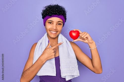 Beautiful african american afro sporty woman doing exercise wearing towel holding heart very happy pointing with hand and finger