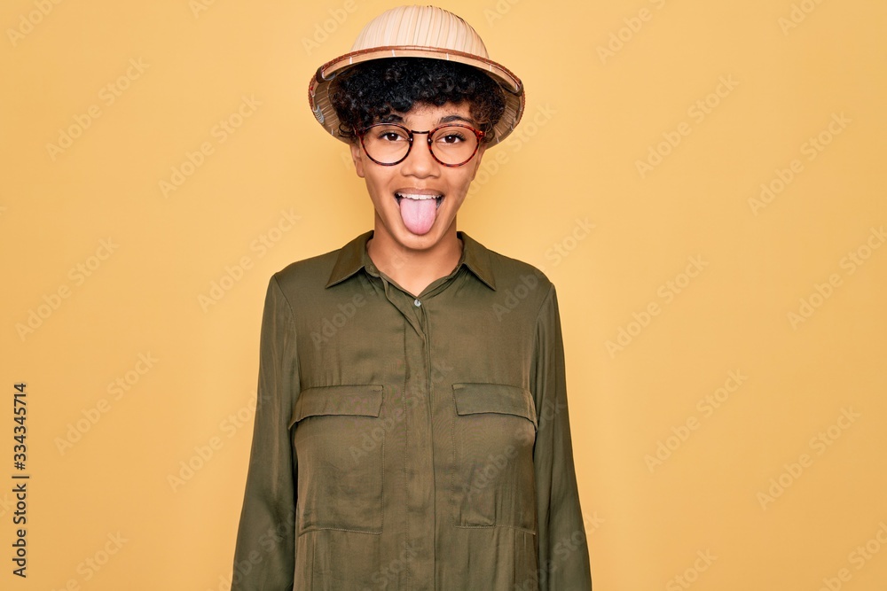 Young beautiful african american tourist woman wearing glasses and explorer hat sticking tongue out happy with funny expression. Emotion concept. Young beautiful african american tourist woman wearing glasses and explorer hat sticking tongue out happy with funny expression. Emotion concept.