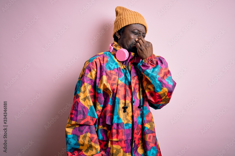 Young handsome african american man wearing colorful coat and cap over pink background smelling something stinky and disgusting, intolerable smell, holding breath with fingers on nose. Bad smell Young handsome african american man wearing colorful coat and cap over pink background smelling something stinky and disgusting, intolerable smell, holding breath with fingers on nose. Bad smell