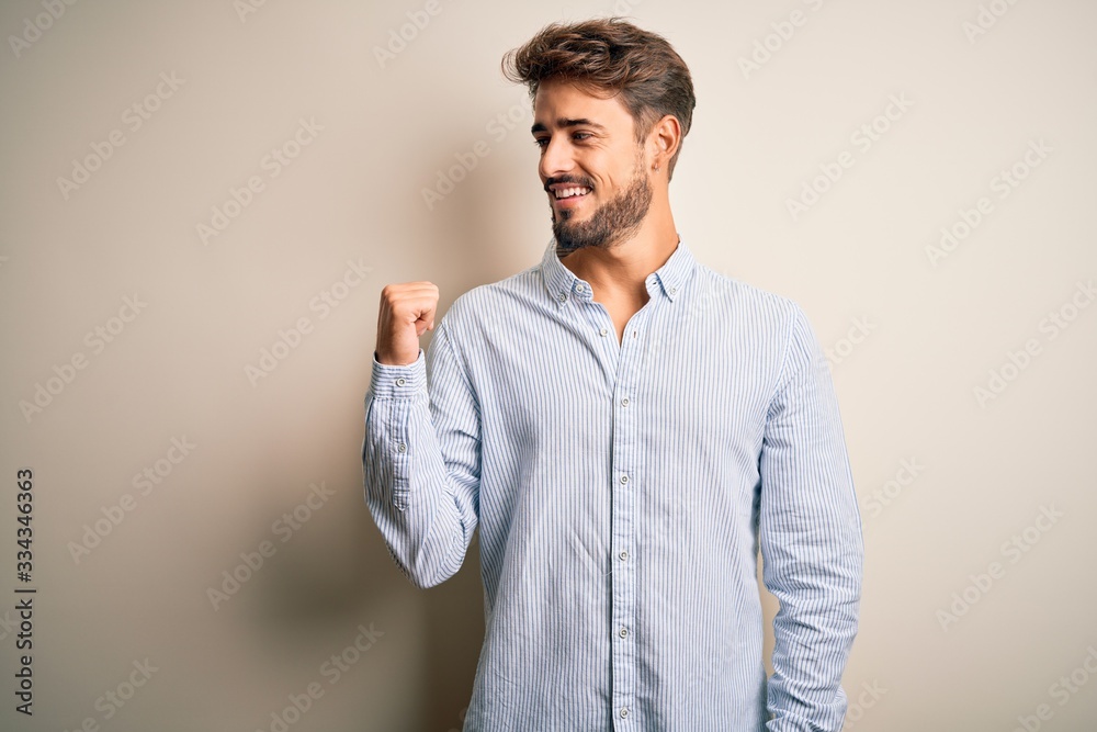 Young handsome man with beard wearing striped shirt standing over white background smiling with happy face looking and pointing to the side with thumb up.