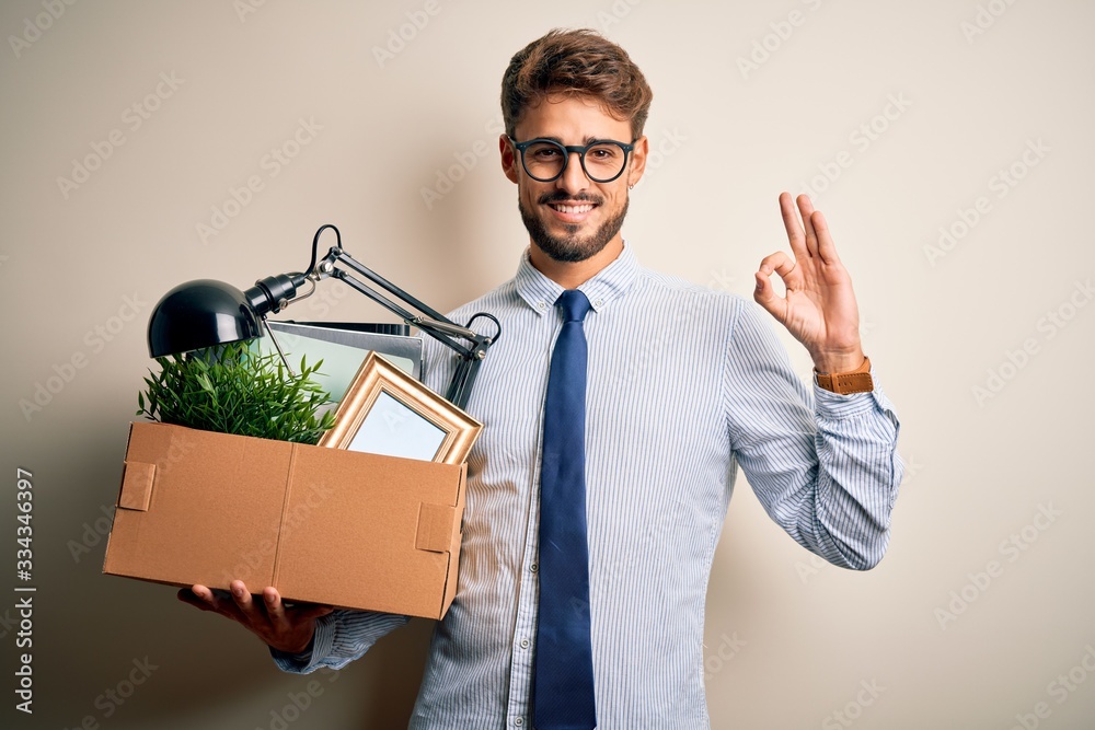 Young businessman with beard fired holding cardboard standing over white background doing ok sign with fingers, excellent symbol Young businessman with beard fired holding cardboard standing over white background doing ok sign with fingers, excellent symbol