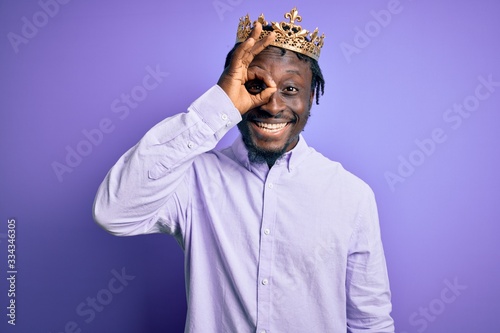 Young african american man wearing golden crown of king over isolated purple background doing ok gesture with hand smiling, eye looking through fingers with happy face.