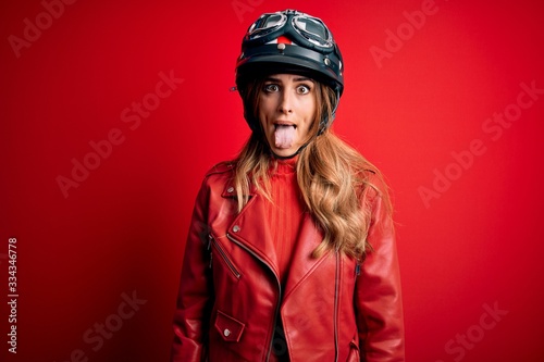 Young beautiful brunette motrocyclist woman wearing moto helmet over red background sticking tongue out happy with funny expression. Emotion concept. © Krakenimages.com