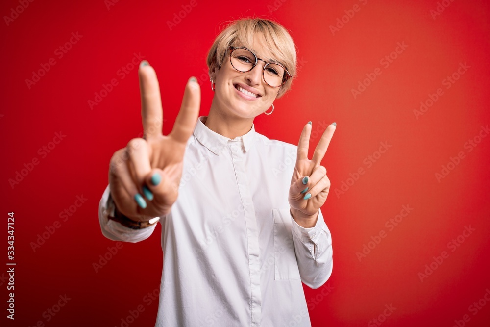 Young blonde business woman with short hair wearing glasses over red background smiling looking to the camera showing fingers doing victory sign. Number two.