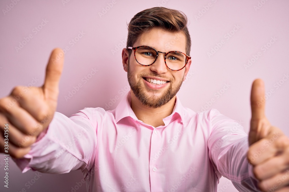 Young handsome blond man with beard and blue eyes wearing pink shirt and glasses approving doing positive gesture with hand, thumbs up smiling and happy for success. Winner gesture. Young handsome blond man with beard and blue eyes wearing pink shirt and glasses approving doing positive gesture with hand, thumbs up smiling and happy for success. Winner gesture.