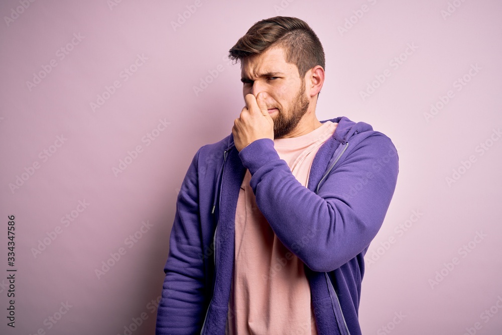 Young blond man with beard and blue eyes wearing purple sweatshirt over pink background smelling something stinky and disgusting, intolerable smell, holding breath with fingers on nose. Bad smell