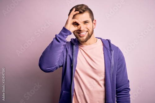 Young blond man with beard and blue eyes wearing purple sweatshirt over pink background doing ok gesture with hand smiling, eye looking through fingers with happy face. © Krakenimages.com