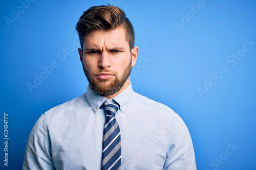 Young blond businessman with beard and blue eyes wearing elegant shirt and tie standing skeptic and nervous, frowning upset because of problem. Negative person.