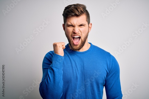 Young handsome blond man with beard and blue eyes wearing casual sweater angry and mad raising fist frustrated and furious while shouting with anger. Rage and aggressive concept.