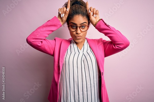 Beautiful african american businesswoman wearing jacket and glasses over pink background doing funny gesture with finger over head as bull horns