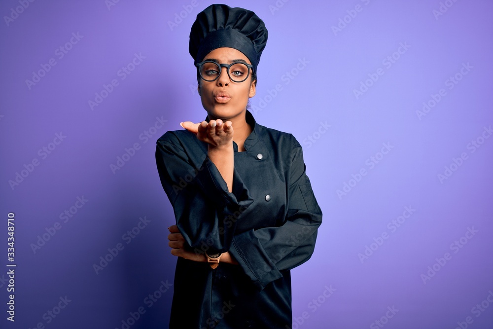 Young african american chef girl wearing cooker uniform and hat over purple background looking at the camera blowing a kiss with hand on air being lovely and sexy. Love expression.