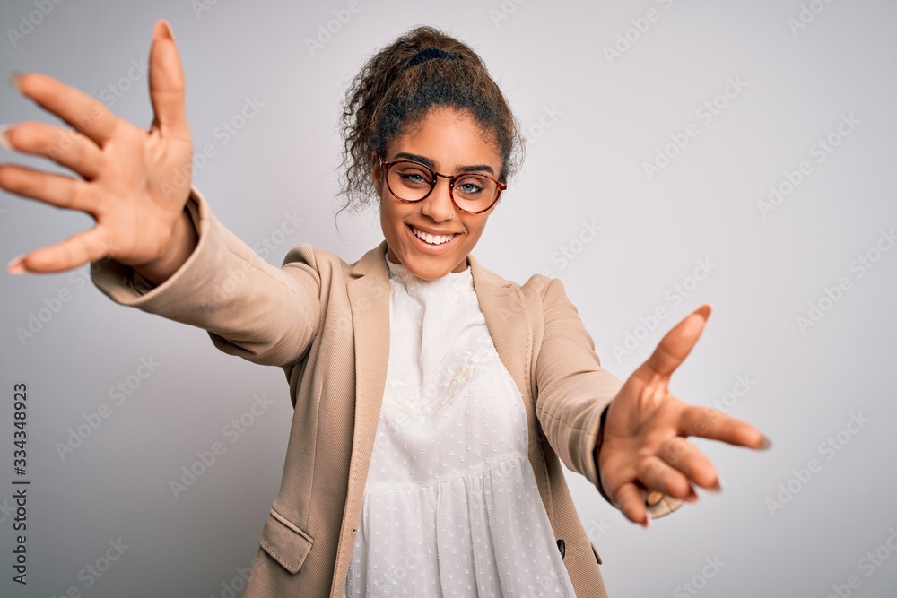 Beautiful african american businesswoman wearing jacket and glasses over white background looking at the camera smiling with open arms for hug. Cheerful expression embracing happiness. Beautiful african american businesswoman wearing jacket and glasses over white background looking at the camera smiling with open arms for hug. Cheerful expression embracing happiness.