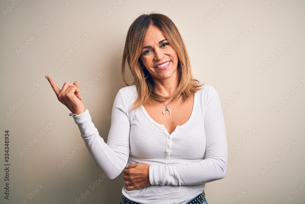 Middle age beautiful woman wearing casual t-shirt standing over isolated white background with a big smile on face, pointing with hand and finger to the side looking at the camera. Middle age beautiful woman wearing casual t-shirt standing over isolated white background with a big smile on face, pointing with hand and finger to the side looking at the camera.