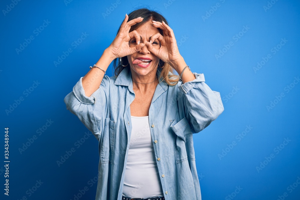 Middle age beautiful woman wearing casual shirt standing over isolated blue background doing ok gesture like binoculars sticking tongue out, eyes looking through fingers. Crazy expression. Middle age beautiful woman wearing casual shirt standing over isolated blue background doing ok gesture like binoculars sticking tongue out, eyes looking through fingers. Crazy expression.
