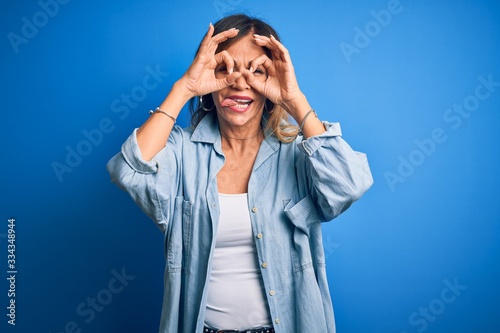 Middle age beautiful woman wearing casual shirt standing over isolated blue background doing ok gesture like binoculars sticking tongue out, eyes looking through fingers. Crazy expression.