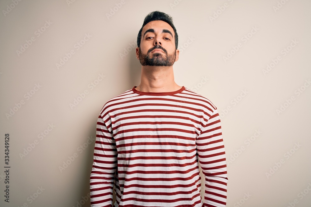 Young handsome man with beard wearing casual striped t-shirt standing over white background Relaxed with serious expression on face. Simple and natural looking at the camera. Young handsome man with beard wearing casual striped t-shirt standing over white background Relaxed with serious expression on face. Simple and natural looking at the camera.
