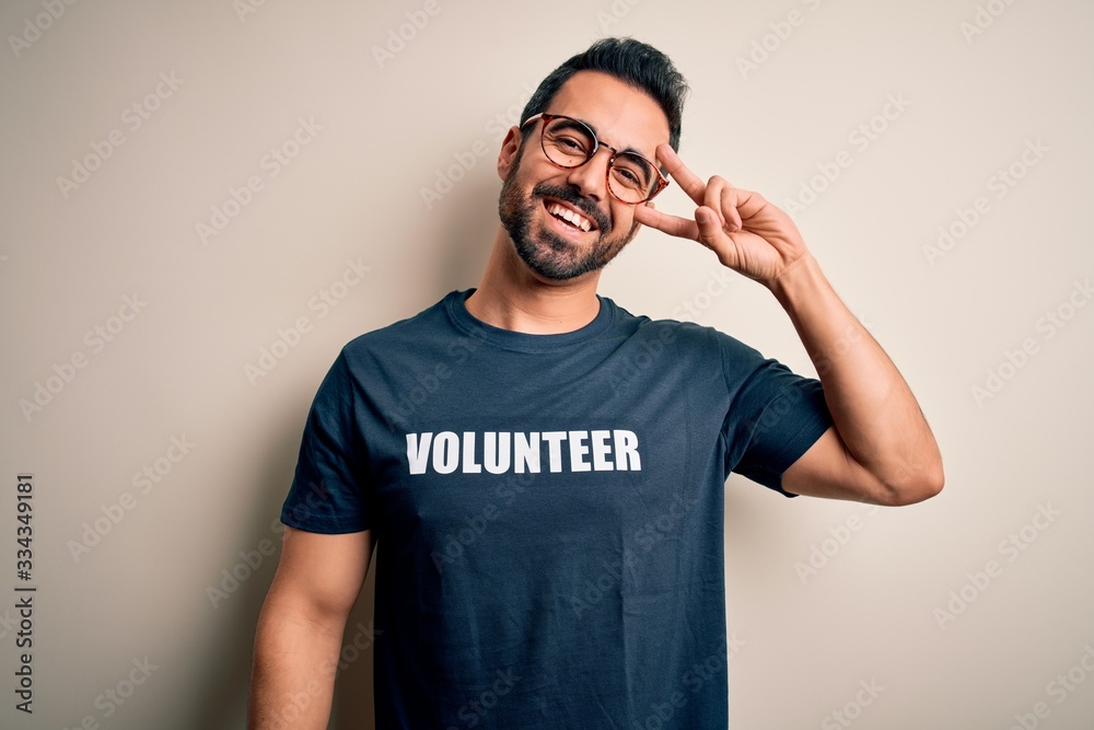 Handsome man with beard wearing t-shirt with volunteer message over white background Doing peace symbol with fingers over face, smiling cheerful showing victory