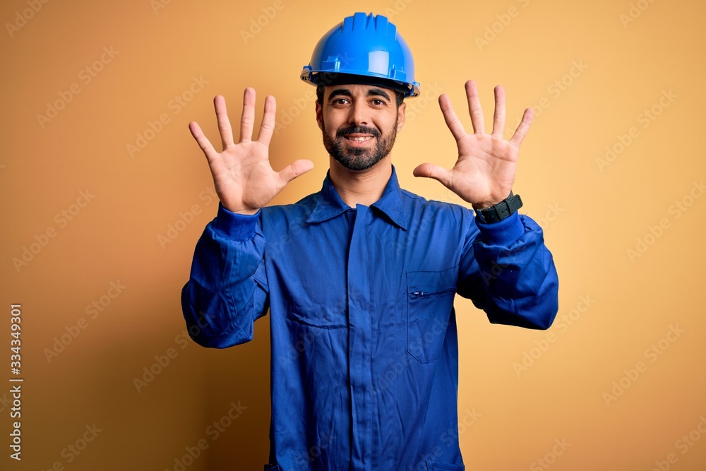 Mechanic man with beard wearing blue uniform and safety helmet over yellow background showing and pointing up with fingers number ten while smiling confident and happy.