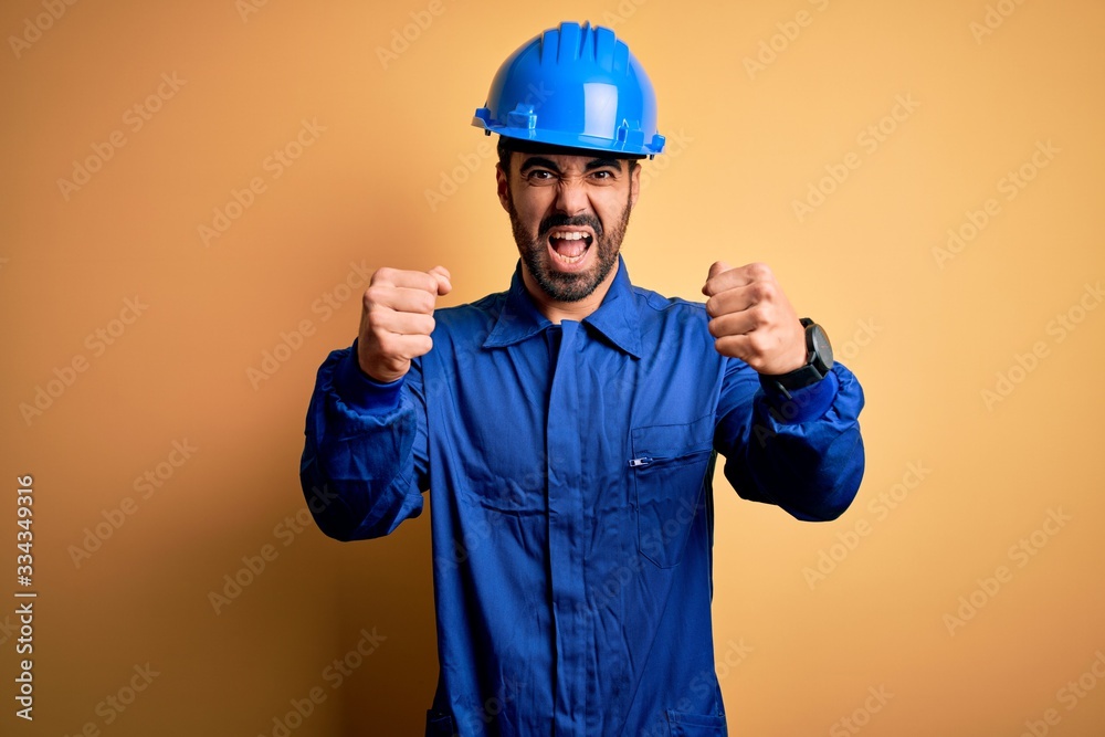 Mechanic man with beard wearing blue uniform and safety helmet over yellow background angry and mad raising fists frustrated and furious while shouting with anger. Rage and aggressive concept.