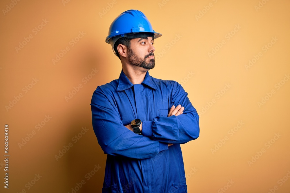 Mechanic man with beard wearing blue uniform and safety helmet over yellow background looking to the side with arms crossed convinced and confident