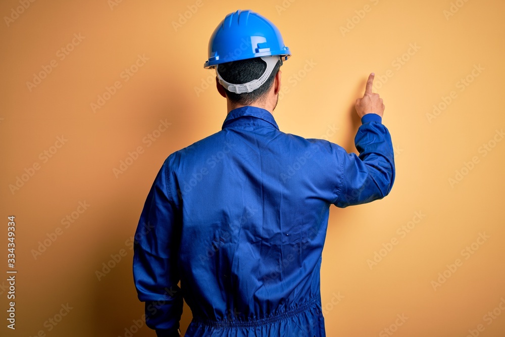 Mechanic man with beard wearing blue uniform and safety helmet over yellow background Posing backwards pointing ahead with finger hand