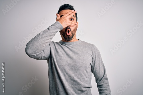 Young handsome man with beard wearing casual sweater standing over white background peeking in shock covering face and eyes with hand, looking through fingers with embarrassed expression.