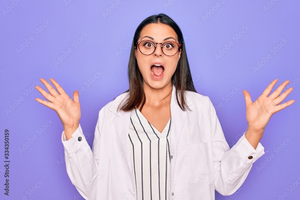 Young beautiful brunette psychologist woman wearing coat and glasses over purple background celebrating crazy and amazed for success with arms raised and open eyes screaming excited. Winner concept Young beautiful brunette psychologist woman wearing coat and glasses over purple background celebrating crazy and amazed for success with arms raised and open eyes screaming excited. Winner concept