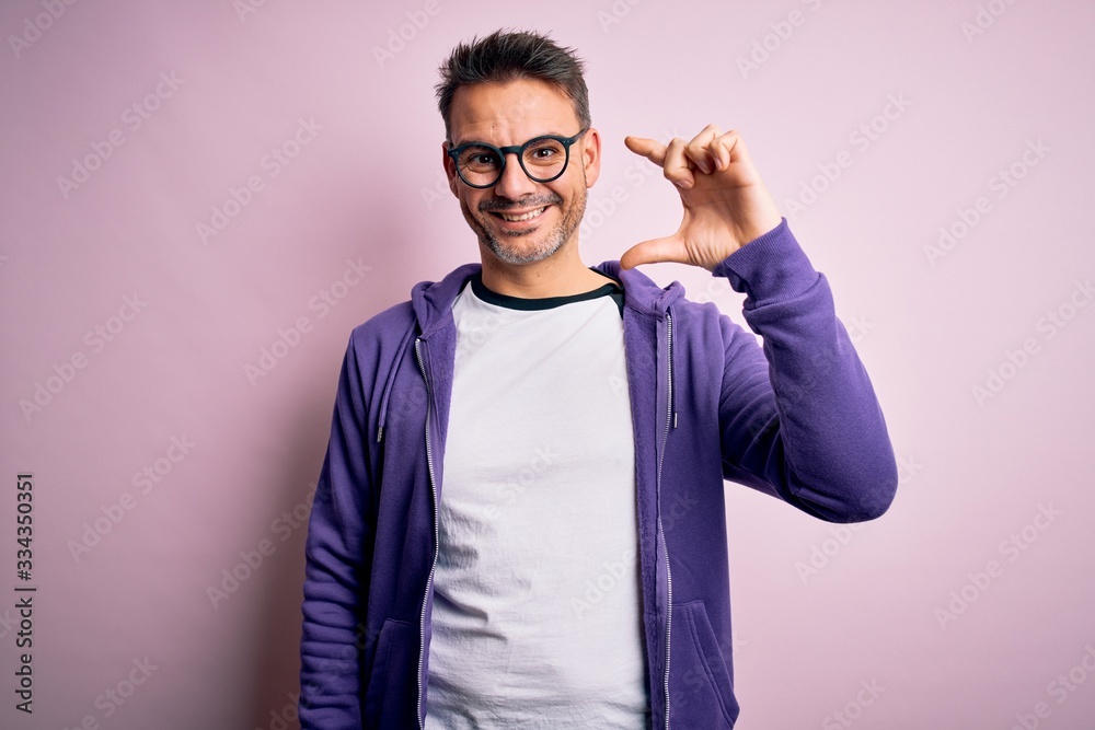 Young handsome man wearing purple sweatshirt and glasses standing over pink background smiling and confident gesturing with hand doing small size sign with fingers looking and the camera. Measure Young handsome man wearing purple sweatshirt and glasses standing over pink background smiling and confident gesturing with hand doing small size sign with fingers looking and the camera. Measure