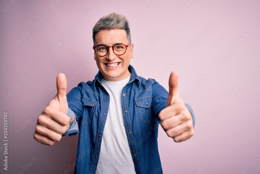 Young handsome modern man wearing glasses and denim jacket over pink isolated background approving doing positive gesture with hand, thumbs up smiling and happy for success. Winner gesture.