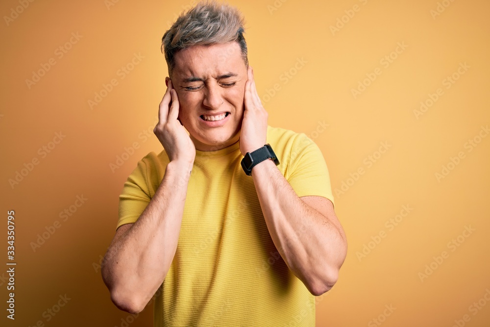 Young handsome modern man wearing yellow shirt over yellow isolated background covering ears with fingers with annoyed expression for the noise of loud music. Deaf concept. Young handsome modern man wearing yellow shirt over yellow isolated background covering ears with fingers with annoyed expression for the noise of loud music. Deaf concept.