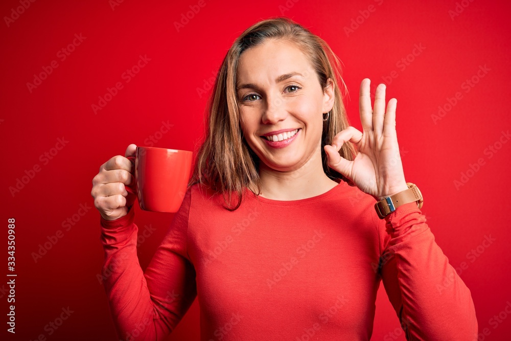 Young beautiful blonde woman drinking cup of coffee standing over isolated red background doing ok sign with fingers, excellent symbol Young beautiful blonde woman drinking cup of coffee standing over isolated red background doing ok sign with fingers, excellent symbol