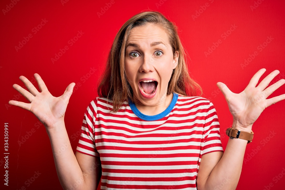 Young beautiful blonde woman wearing casual striped t-shirt over isolated red background celebrating crazy and amazed for success with arms raised and open eyes screaming excited. Winner concept Young beautiful blonde woman wearing casual striped t-shirt over isolated red background celebrating crazy and amazed for success with arms raised and open eyes screaming excited. Winner concept